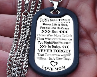 May include: A black dog tag pendant with a silver chain. The tag has engraved text including "To My Son STEVEN" and "LOVE MOM". The chain is a curb style, and the pendant is held in a hand.