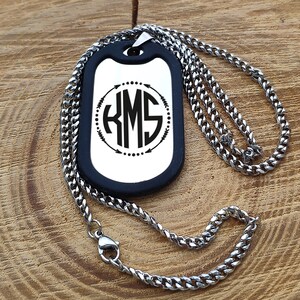 May include: A silver chain necklace with a black dog tag pendant. The dog tag has a silver background with a black circle containing the monogram "KMS" surrounded by arrows and dots.