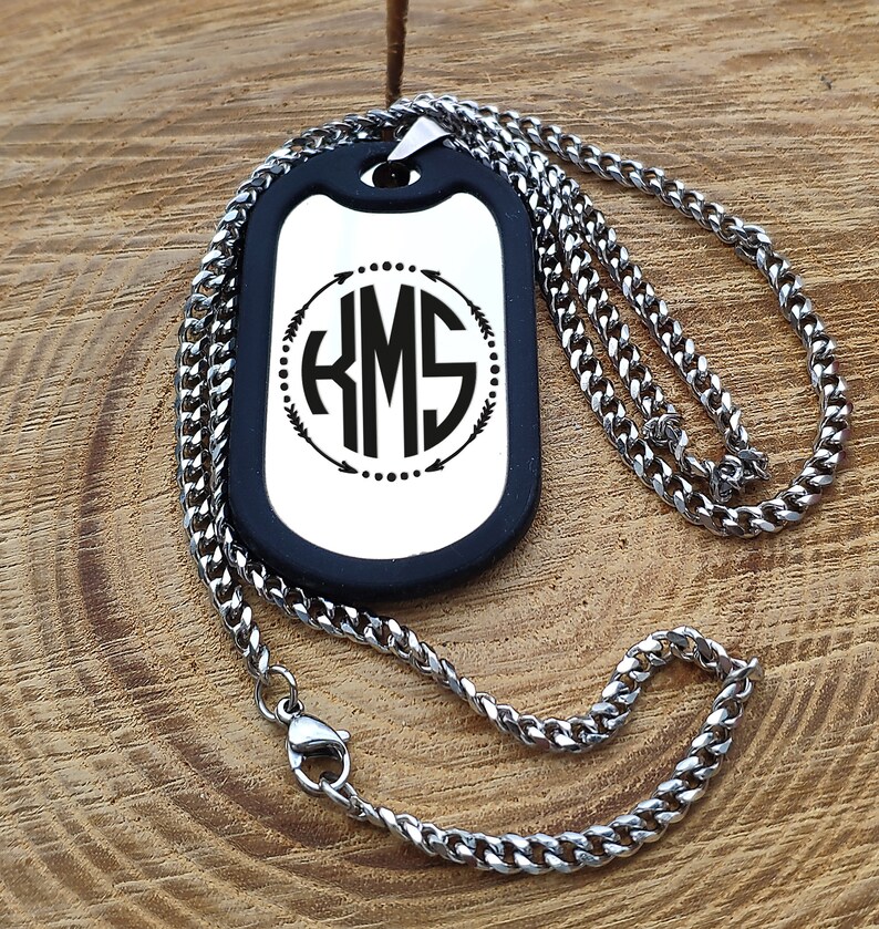 May include: A silver chain necklace with a black dog tag pendant. The dog tag has a silver background with a black circle containing the monogram "KMS" surrounded by arrows and dots.