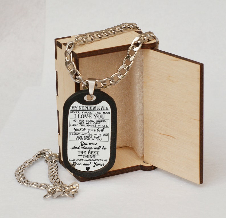 May include: A black dog tag necklace with a silver chain in a wooden box. The dog tag has an inscription that reads "My Nephew Kyle Never forget how much I love you As you grow older you will face many challenges in life Just do your best I might not be with you but know that I believe in you You were and always will be the best -thing- that ever happened to me Love, aunt James"
