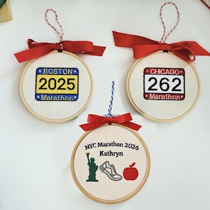 May include: Three round wooden embroidery hoop ornaments with fabric centers. One features a blue and yellow Boston 2025 Marathon design, one a red and white Chicago 262 Marathon design, and one an NYC Marathon 2026 design with a Statue of Liberty, shoe, and apple.