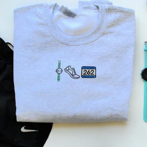 May include: A gray sweatshirt with an embroidered design of a watch, running shoe, and a race bib with the number 262.