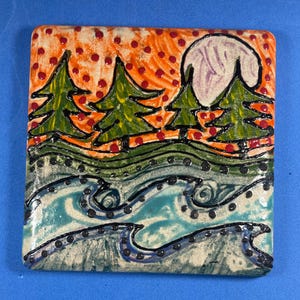 May include: A hand-painted ceramic tile with a whimsical design featuring a sunset with orange and red dots, four green trees, and a purple moon. The bottom of the tile depicts a blue and green river with black swirls and dots.