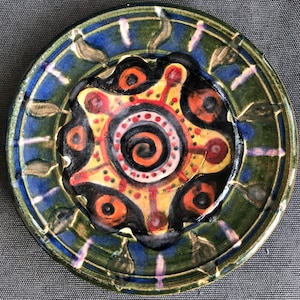 Puede incluir: Un plato de cerámica con un diseño verde, azul y amarillo. El centro del plato presenta un sol estilizado con una espiral en el medio.