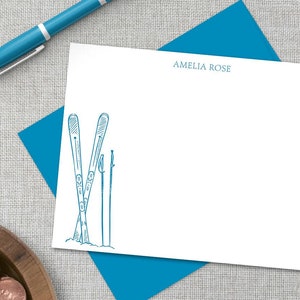 May include: A white card with blue text and a blue envelope. The card has a drawing of skis and ski poles. The text on the card reads "Amelia Rose".