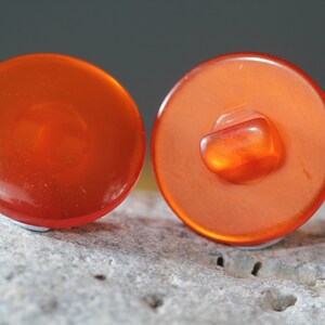 Op de afbeelding: Twee oranje, ronde knopen met een glanzende afwerking. E&eacute;n knoop toont de achterkant met een vierkante schacht. De knopen liggen op een getextureerd, lichtgrijs oppervlak.