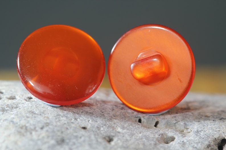 Op de afbeelding: Twee oranje, ronde knopen met een glanzende afwerking. E&eacute;n knoop toont de achterkant met een vierkante schacht. De knopen liggen op een getextureerd, lichtgrijs oppervlak.