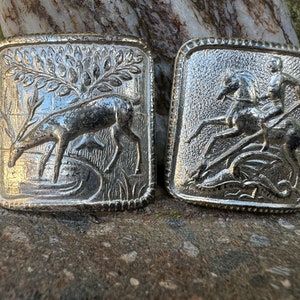 May include: Two silver belt buckles with detailed engravings. One buckle features a deer drinking from a stream, while the other depicts a knight on horseback slaying a dragon.