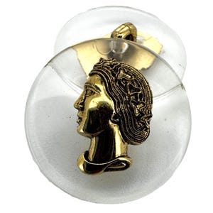 May include: A vintage button featuring a gold-toned profile of a woman with flowing hair, set against a clear, circular base. The button has a gold-toned shank on the back. The button is approximately 1.5 inches in diameter.