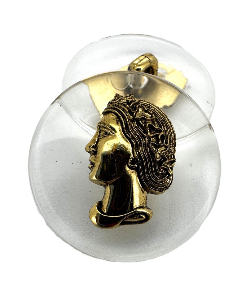 May include: A vintage button featuring a gold-toned profile of a woman with flowing hair, set against a clear, circular base. The button has a gold-toned shank on the back. The button is approximately 1.5 inches in diameter.