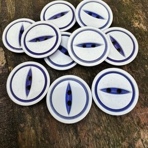 May include: A collection of ten white ceramic buttons with a blue rim and a blue eye-shaped design. Each button has three holes.
