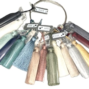 May include: A selection of decorative tassels in shades like black, white, blue, green, and blush. Each tassel has a small tag with a colour name. The tassels are displayed on a silver metal ring.