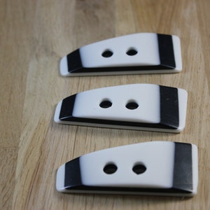 May include: Three black and white plastic buttons with two holes each. The buttons are shaped like a rounded rectangle with a black stripe on the top and bottom.