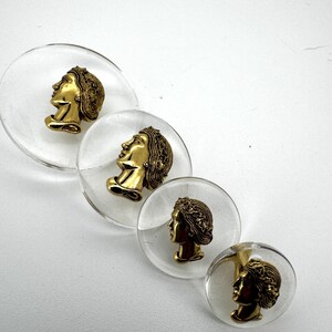 May include: Four clear, circular decorative buttons with gold-colored profile busts encased within. The busts feature detailed hair and facial features. The buttons are arranged diagonally against a white background.
