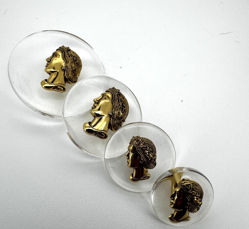 May include: Four clear, circular decorative buttons with gold-colored profile busts encased within. The busts feature detailed hair and facial features. The buttons are arranged diagonally against a white background.