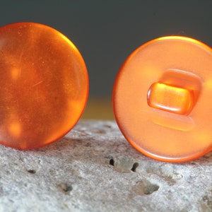 Op de afbeelding: Twee oranje, ronde knopen met een glanzende afwerking. E&eacute;n knoop toont de voorkant, de andere de achterkant met een rechthoekige schacht. De knopen staan tegen een neutrale achtergrond.