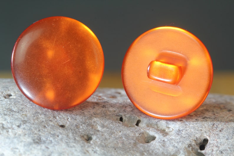 Op de afbeelding: Twee oranje, ronde knopen met een glanzende afwerking. E&eacute;n knoop toont de voorkant, de andere de achterkant met een rechthoekige schacht. De knopen staan tegen een neutrale achtergrond.
