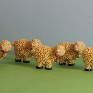 Può includere: Cinque piccole statuine di pecore bianche e soffici con campanelle rosse e dorate intorno al collo. Le pecore sono in piedi su una superficie verde.