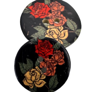 Könnte beinhalten: Zwei runde Holzknöpfe mit einem floralen Rosendesign. Die Knöpfe sind schwarz mit roten, orangefarbenen und gelben Rosen und grünen Blättern. Jeder Knopf hat zwei Löcher zum Nähen.