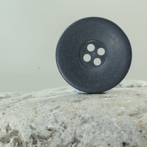May include: A large, dark gray button with four white holes in the center. The button is round and sits on a gray, textured rock. The button is a sewing or craft supply.