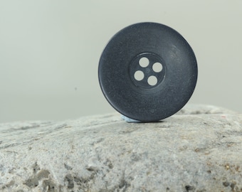 May include: A large, dark gray button with four white holes in the center. The button is round and sits on a gray, textured rock. The button is a sewing or craft supply.
