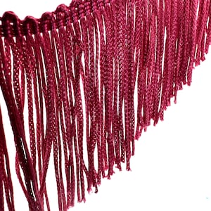 May include: A close-up of a burgundy fringe trim with a woven edge. The fringe is made of thin, soft fibres and is approximately 2.5 cm in length.