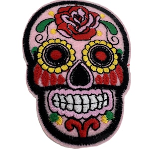 May include: Embroidered patch in the shape of a sugar skull, with a pink background and black outlining. The skull has yellow sunflower eyes, a red rose on top, and a white toothy grin. The design is a colourful representation of the Day of the Dead.