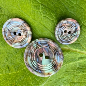 May include: Three round buttons with a marbled pattern in shades of blue, green, and brown. The buttons have two holes each and a raised edge.