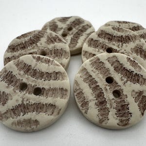 Può includere: Un set di sei bottoni in ceramica con un motivo a spirale marrone e bianco. I bottoni hanno due fori ciascuno.