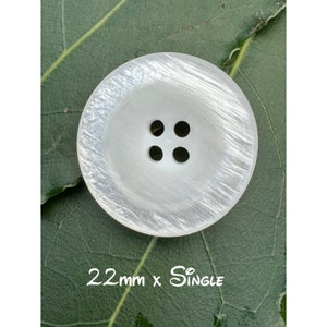 May include: A large, round, pearlescent white button with four holes. The button has a subtle, textured pattern and is 22mm in diameter. The text "22mm x Single" is visible at the bottom of the image. The button is set against a green leaf.