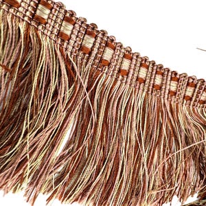 May include: Close-up of decorative fringe trim. The trim features a woven top section in shades of brown and beige, with a fringe of long, thin threads in brown, beige, and pink hues. The background is white.