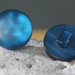 Op de afbeelding: Twee ronde, blauwgroene knopen. E&eacute;n knoop ligt met de voorkant naar boven en toont een glad, enigszins doorschijnend oppervlak. De andere knoop ligt met de achterkant naar beneden en onthult een schacht om te naaien. De knopen liggen op een getextureerd, grijs oppervlak.