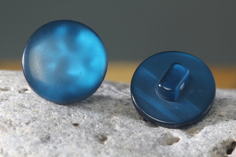 Op de afbeelding: Twee ronde, blauwgroene knopen. E&eacute;n knoop ligt met de voorkant naar boven en toont een glad, enigszins doorschijnend oppervlak. De andere knoop ligt met de achterkant naar beneden en onthult een schacht om te naaien. De knopen liggen op een getextureerd, grijs oppervlak.
