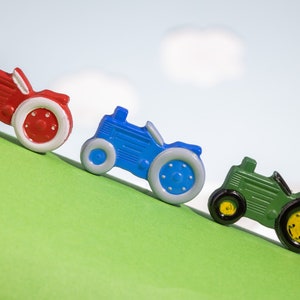 May include: Three toy tractors lined up on a green surface. The tractors are red, blue, and green. The red tractor is at the front, followed by the blue tractor, and then the green tractor.