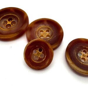 Könnte beinhalten: Vier braune Plastikknöpfe mit jeweils vier Löchern. Die Knöpfe sind in verschiedenen Größen und haben eine braune Marmorierung.