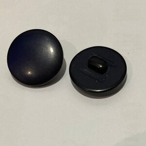 Op de afbeelding: Twee marineblauwe ronde knopen. E&eacute;n knoop ligt met de voorkant naar boven en toont een glad, licht reflecterend oppervlak. De andere knoop ligt met de achterkant naar beneden en onthult een zwarte schacht. Deze knopen zijn geschikt voor diverse naaiprojecten.