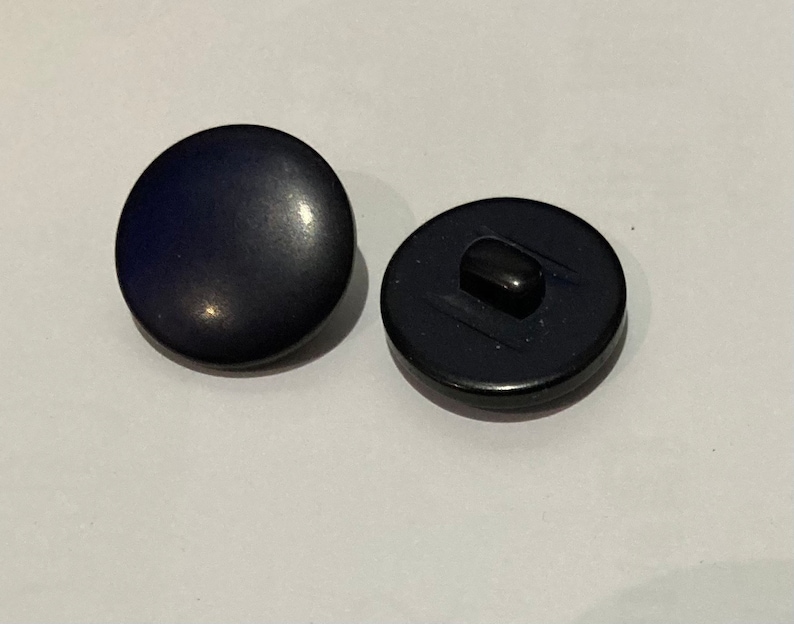 Op de afbeelding: Twee marineblauwe ronde knopen. E&eacute;n knoop ligt met de voorkant naar boven en toont een glad, licht reflecterend oppervlak. De andere knoop ligt met de achterkant naar beneden en onthult een zwarte schacht. Deze knopen zijn geschikt voor diverse naaiprojecten.