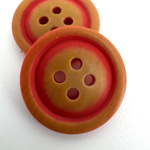 May include: Two round buttons with a wood-grain effect. Each button has a reddish-orange centre ring and four holes. The buttons are a warm brown colour with a reddish-orange accent.