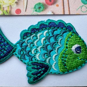 May include: An embroidered fish patch in blue and green with white details. The fish has a large eye and a scalloped fin.