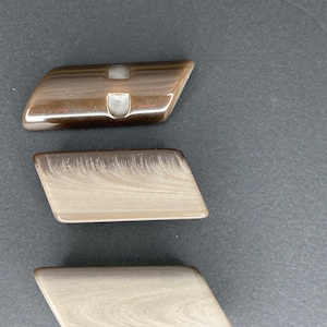 Op de afbeelding: Drie bruine en witte plastic knoppen met een gladde, glanzende afwerking. De knoppen hebben de vorm van een rechthoek met afgeronde hoeken. De bovenste knop heeft een klein gaatje in het midden.