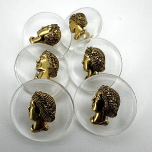 May include: Clear, round cabinet knobs with a gold-colored profile of a woman's head. The knobs are set against a white background. The woman's head has detailed hair and facial features. The knobs are likely made of glass or acrylic.