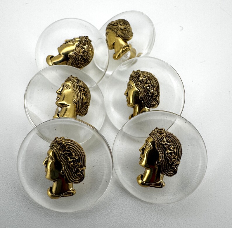 May include: Clear, round cabinet knobs with a gold-colored profile of a woman's head. The knobs are set against a white background. The woman's head has detailed hair and facial features. The knobs are likely made of glass or acrylic.