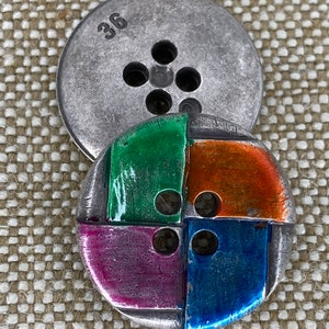 May include: A silver metal button with four holes and the number 36 printed on it. The button is layered on top of a colourful button with four sections of green, orange, purple, and blue enamel.