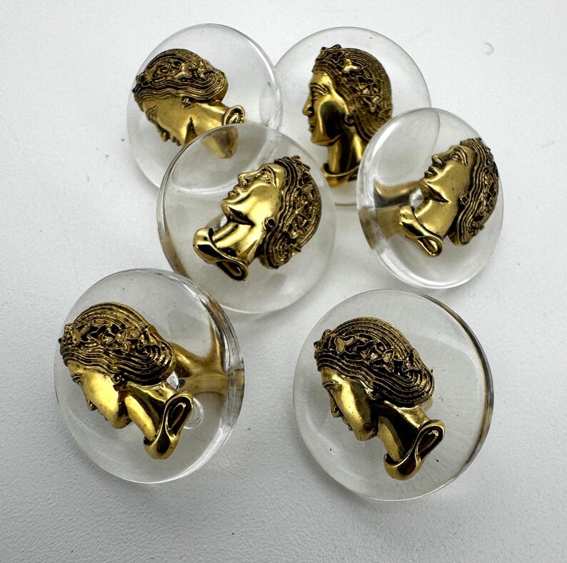 May include: Six clear, round buttons with a gold-colored profile of a woman's face. The profile is detailed with flowing hair and a classical design. The buttons are set against a white background.