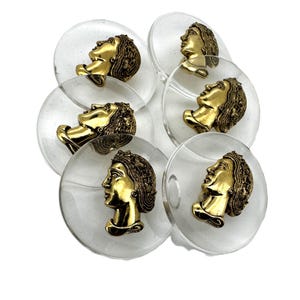 May include: A collection of clear, circular buttons with a gold-colored profile of a person's face. The buttons are arranged in a slightly overlapping pattern against a white background. The face profiles are detailed and ornate.