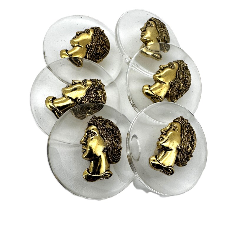 May include: A collection of clear, circular buttons with a gold-colored profile of a person's face. The buttons are arranged in a slightly overlapping pattern against a white background. The face profiles are detailed and ornate.