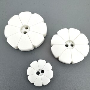 May include: Three white flower-shaped buttons in different sizes. Each button features a petal design and two holes in the centre for sewing. The buttons are arranged on a grey surface.