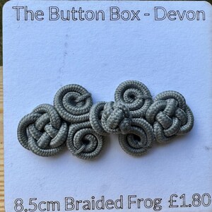 May include: A gray braided frog closure, 8.5 inches long, for clothing or crafts. The button is made of woven cord and has a knot design. The Button Box - Devon is printed on the white background.