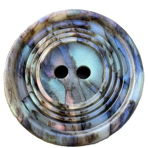 May include: A round, two-hole button featuring a swirling, marbled pattern in shades of iridescent blue, teal, purple, and cream. The button has a recessed centre and a raised, smooth outer rim, creating a layered, circular design with a glossy finish.