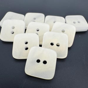 May include: A group of ten white, square buttons with two holes each. The buttons have a slightly iridescent sheen.